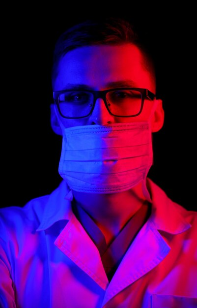 Doctor wears mask. Black background with blue - red light. Doctor in glasses with medical mask on face. Close up shot. Virus, COVID-19 and coronavirus identification and prevention.