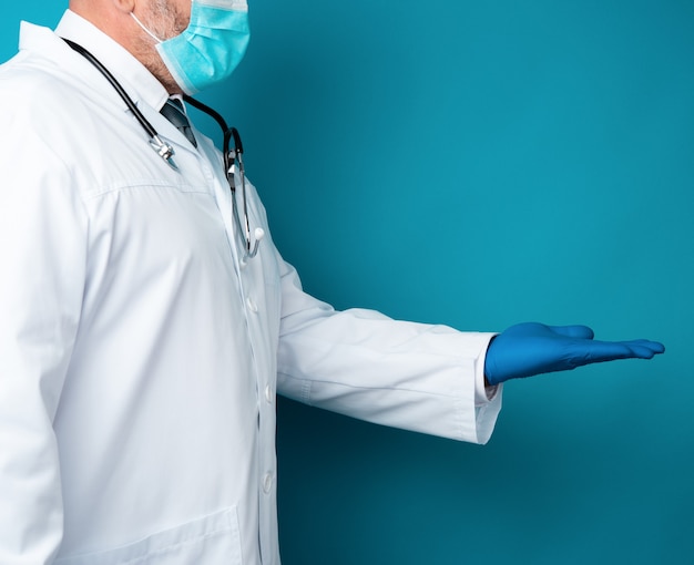 Doctor in a white coat, blue latex gloves and a protective mask on his face conditionally holds an object with his hand on a blue background