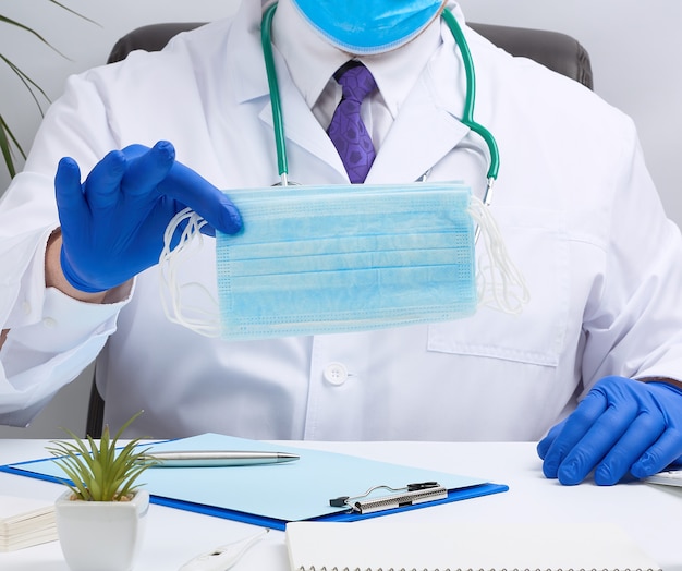 Doctor in a white coat and blue latex gloves sits at a white desk and shows a stack of disposable medical masks