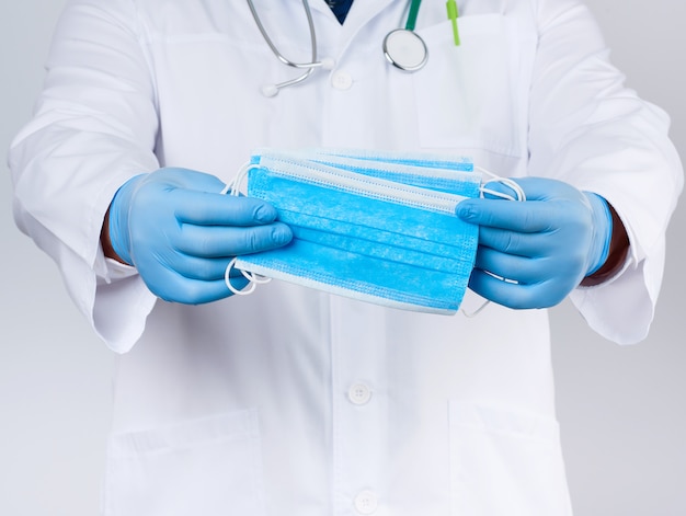 Doctor in a white coat, blue latex sterile gloves holds textile medical masks in his hand