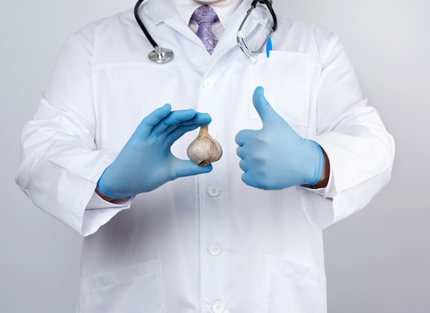 doctor in a white medical coat and blue rubber gloves holds a fresh head of garlic, prevention against viral diseases