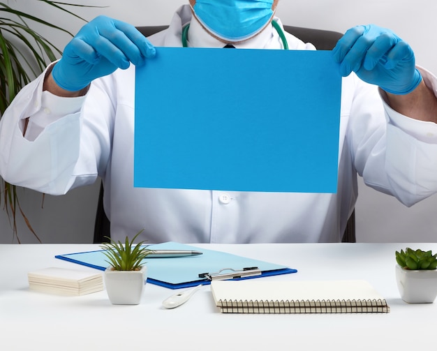 Doctor in a white medical coat is sitting at a table in a brown leather chair and holding a empty blue sheet of paper in his hands, place for an inscription, medical office