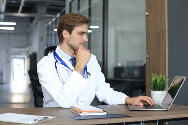 Doctor in white medical uniform with statoscope reading personal file of patient on tablet and writes data to laptop.