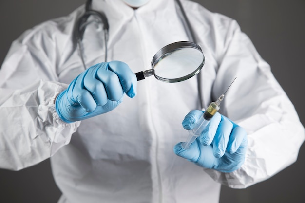 Doctor in white protective uniform examining vaccine with magnifier on gray scene