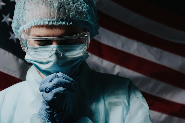 Photo a doctor who is tired after a hard day, the doctor prays against the backdrop of the american flag and asks god for help and strength in the fight against the coronavirus epidemic