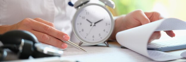 Doctor with alarm clock and documents on hospital table female cardiologist looking at medical