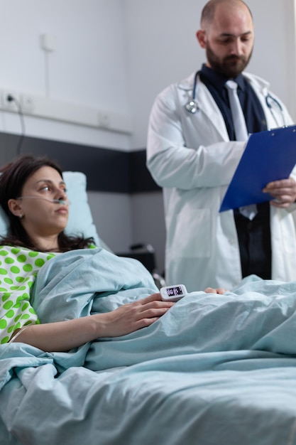 Doctor with clipboard reading medical data to patient with nasal cannula recieving oxygen in hospital bed. Patient having lung disease with pulse oximeter on finger showing low spo2 saturation.