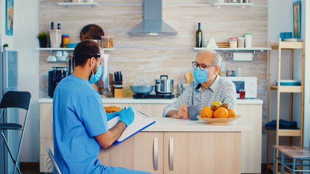 Doctor with face mask and visor writing prescription on clipboard during patient home visit. Male nurse social worker at retired senior couple visit explaining covid-19 spreading, help for people in r