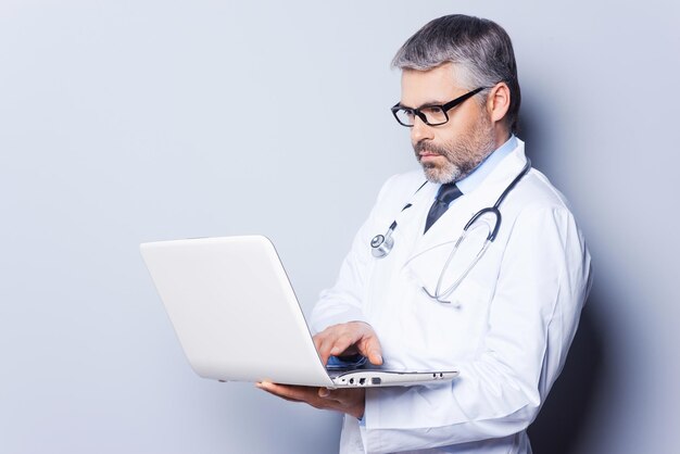 Doctor with laptop. Confident mature doctor working on laptop while standing against grey background