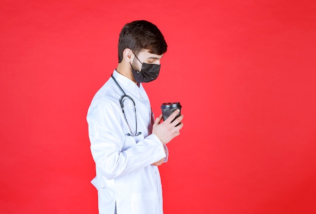 Doctor with stethoscope in black mask holding a black takeaway coffee cup. 