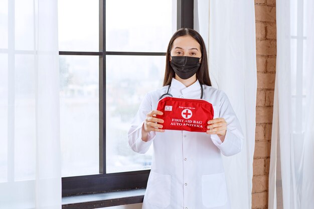 Doctor with stethoscope and black mask holding a red first aid kit. 