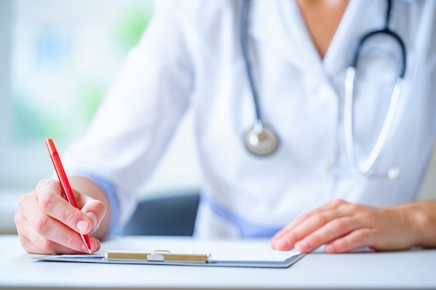 Doctor with a stethoscope writing diagnosis and therapy of the disease in a clipboard during a medical consultation in hospital. Healthcare and medical prescription for pills and medicine