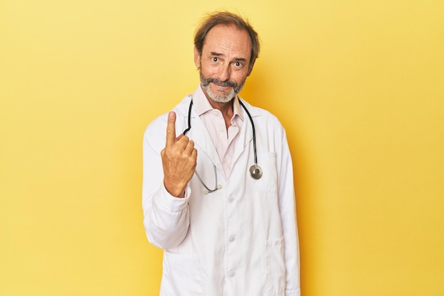 Doctor with stethoscope in yellow studio pointing with finger at you as if inviting come closer