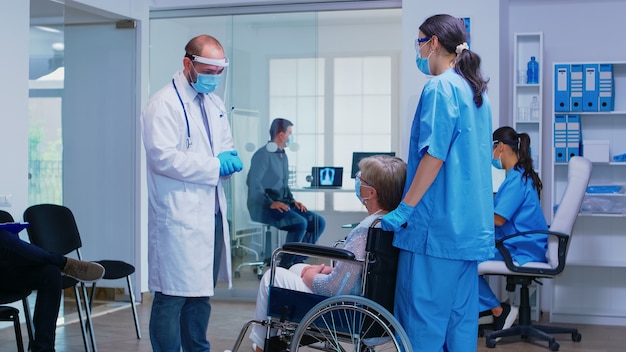 Doctor with visor against coronavirus discussing with disabled senior woman in wheelchair in hospital waiting area and patient is waiting in examination room.