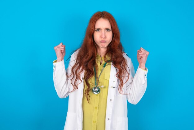 Doctor woman blows cheeks with anger and raises clenched fists expresses rage and aggressive