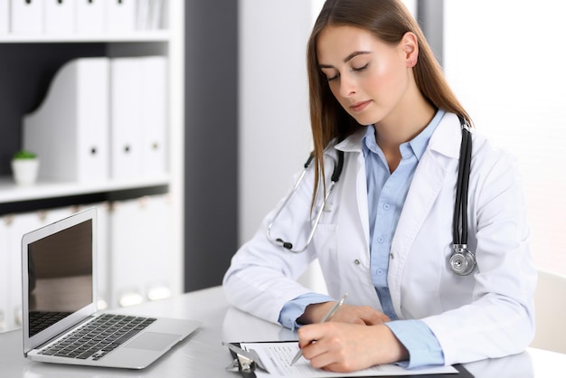 Женщина-врач заполняет медицинскую форму, сидя за столом в больничном кабинете. Врач за работой. Концепция медицины и здравоохранения.