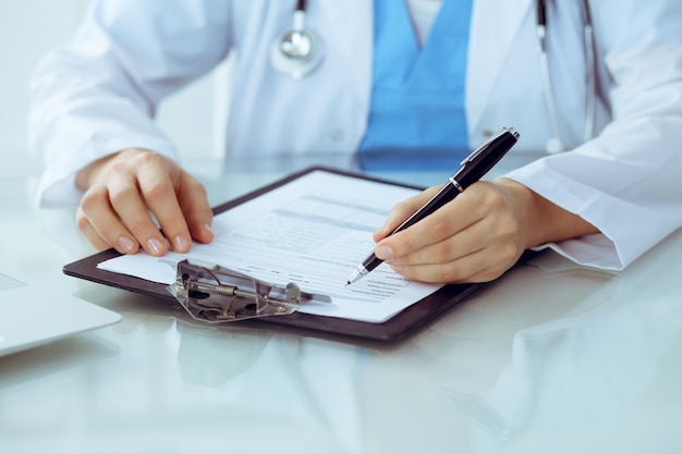 Doctor woman filling up medical form while sitting at the table closeup of hands