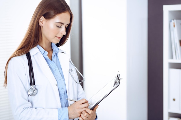 Doctor woman filling up medical form while standing near window in clinic. Physician at work. Medicine and health care concept.