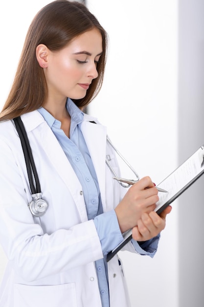 Doctor woman filling up medical form while standing near window in hospital office. Happy physician at work. Medicine and health care concept.