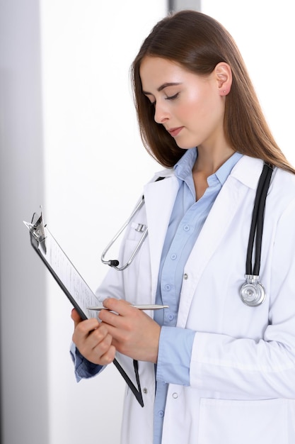 Doctor woman filling up medical form while standing near window in hospital office. Happy physician at work. Medicine and health care concept.