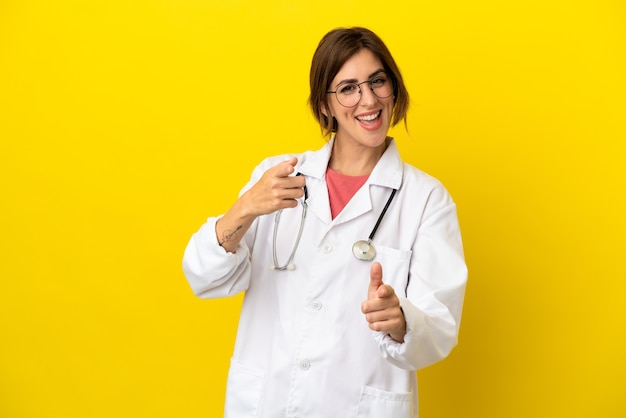 Donna medico isolata su sfondo giallo che punta verso la parte anteriore e sorridente