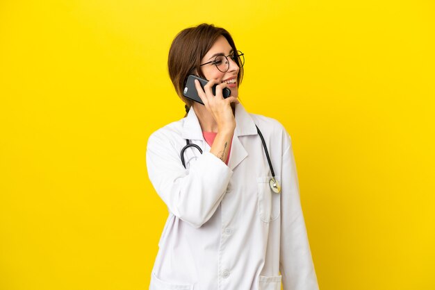 Doctor woman isolated on yellow wall keeping a conversation with the mobile phone with someone