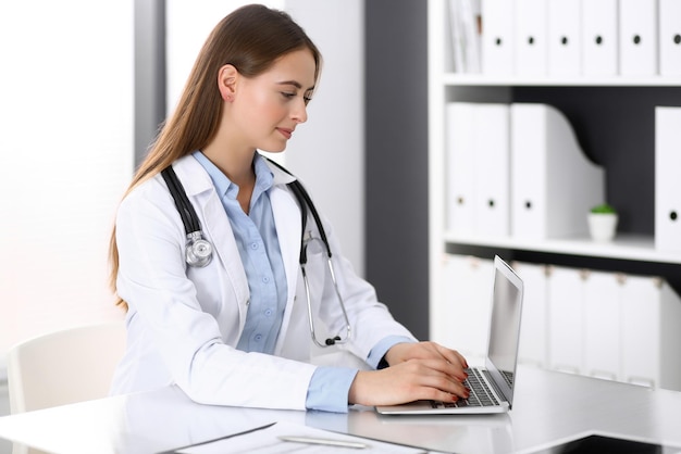 Doctor woman typing on laptop computer while sitting at the desk in hospital office. Physician at work. Medicine and health care concept.