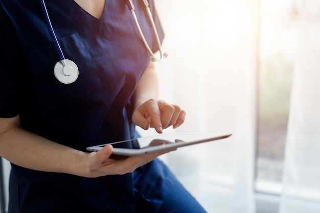 Doctor woman using tablet computer while standing near panorama window in clinic, close up. Physician or surgeon at work. Medicine concept.