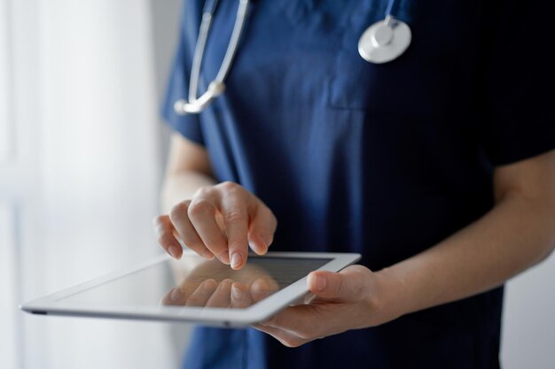 Doctor woman using tablet computer while standing near panorama window in clinic close up physician or surgeon at work medicine concept