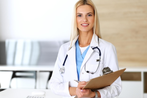 Doctor woman at work in hospital excited and happy of her profession Blonde physician controls medication history records and exam results while using clipboard Medicine and healthcare concept