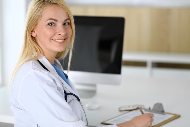 Doctor woman at work while sitting at the desk in hospital or\
clinic blonde cheerful physician ready to help patients data and\
best service in medicine and healthcare