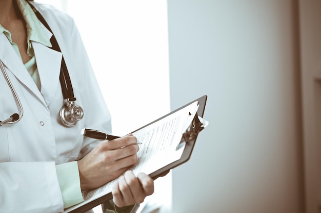 Doctor woman working with medication history records form while standing near window in hospital. Medicine and health care concept. Green is main color.