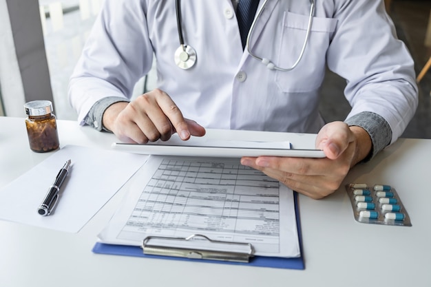 写真 病院や医療聴診器での紙のレポート、机のクリップボードにある薬のデジタルタブレットノート情報を使用して作業している医師。