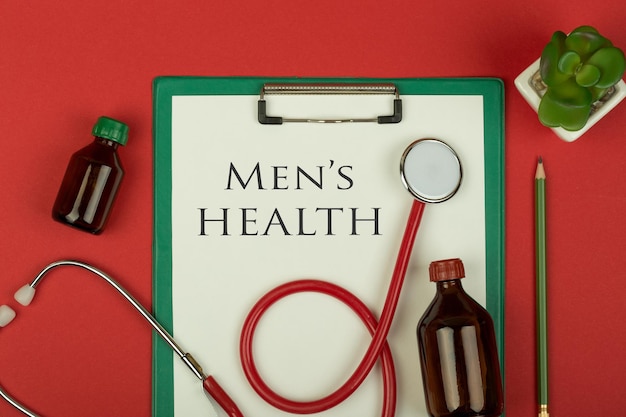 Doctor workplace red stethoscope medical bottles and green clipboard with text Men's health on red paper background