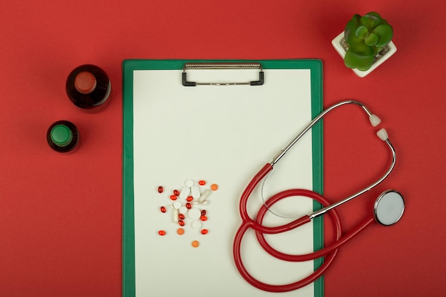 Doctor workplace red stethoscope pills medical bottles and empty green clipboard on red