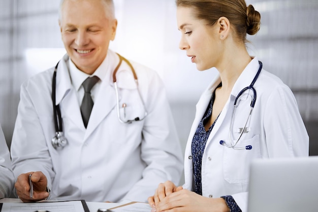 Doctors colleagues and elderly therapist discussing current disease therapy while sitting at working place in sunny clinic. Teamwork in medicine.