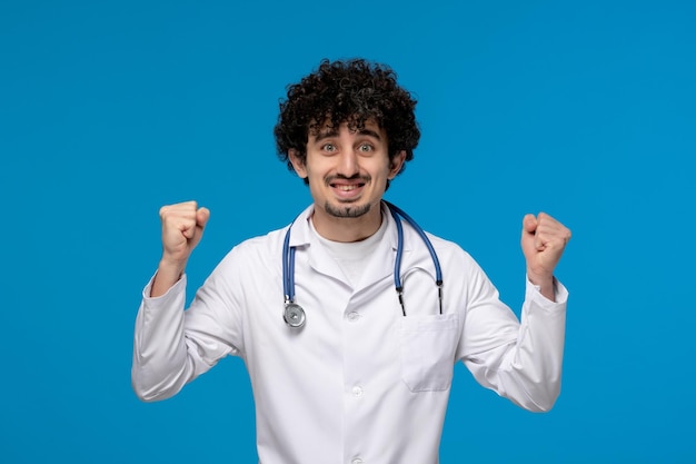 Dottori giorno ricci bel ragazzo carino in uniforme medica che tiene eccitantemente i pugni in alto