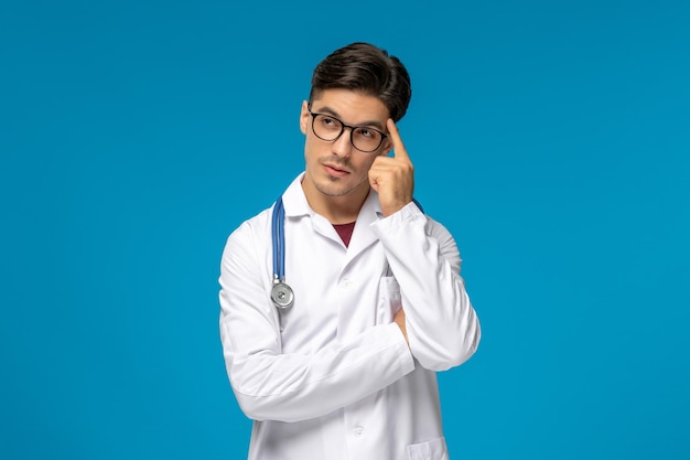 Doctors day cute young brunette guy in lab coat wearing glasses with phonendoscope thinking