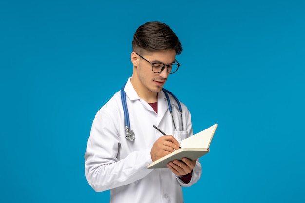 Doctors day cute young handsome man in lab coat and glasses writing down in notebook