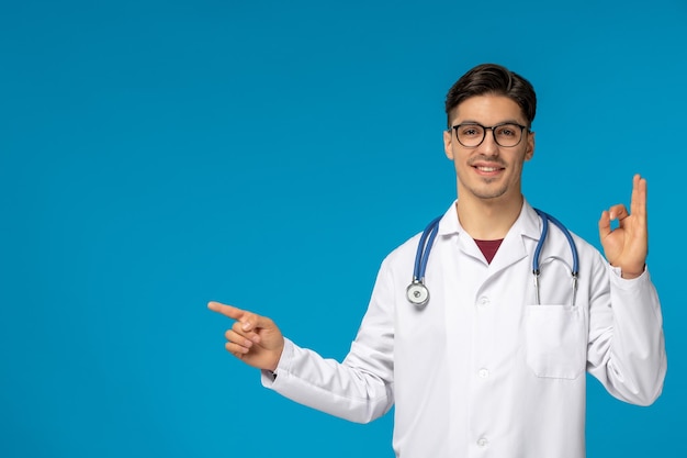 Doctors day handsome brunette cute guy in medical gown showing ok gesture and smiling