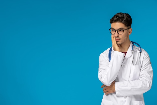 Photo doctors day handsome brunette cute guy in medical gown wearing glasses and thinking