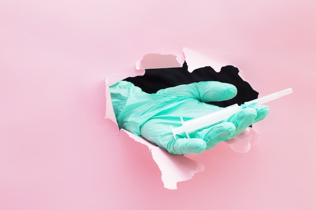 A doctors hand holds out a syringe through a hole in torn paper on a pink background