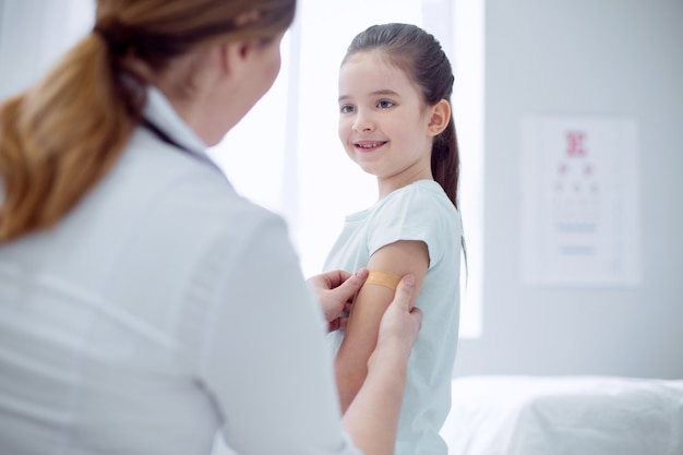 Doctors treatment. Jolly merry girl standing and smiling while doctor pressing patch against her arm