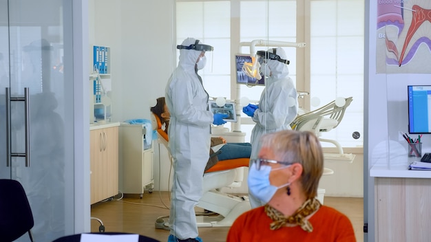 Doctors wearing full virus protection uniform standing in surgery room planning dental treatment while elderly patients waiting in reception keeping distance. Concept of new normal dentist visit