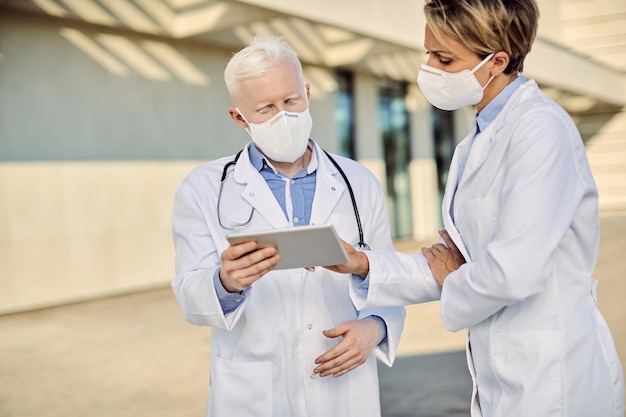 Doctors with protective face masks cooperating while using touchpad outdoors