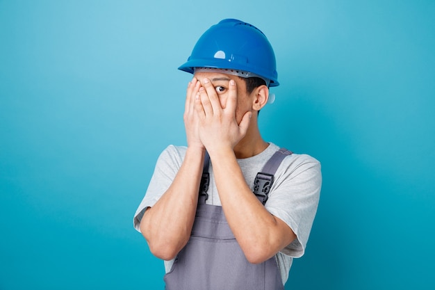 Doen schrikken jonge bouwvakker die veiligheidshelm en uniform bedekkend gezicht draagt met handen die naar camera tussen vingers gluren