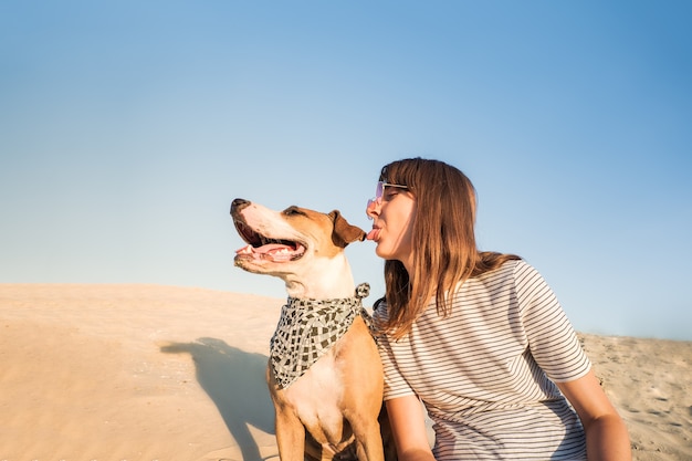 Dog and human make fun, posing as best friends. Funny female person and staffordshire terrier puppy sit on sand on hot summer day