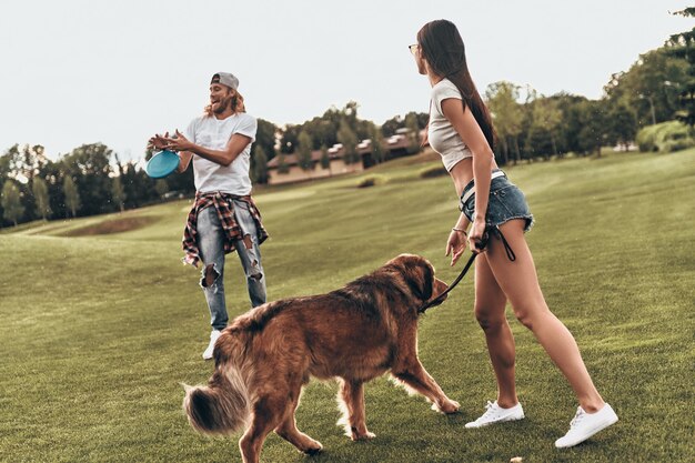 Dog keeps them active.  Full length of beautiful young couple throwing plastic disc while playing with their dog outdoors