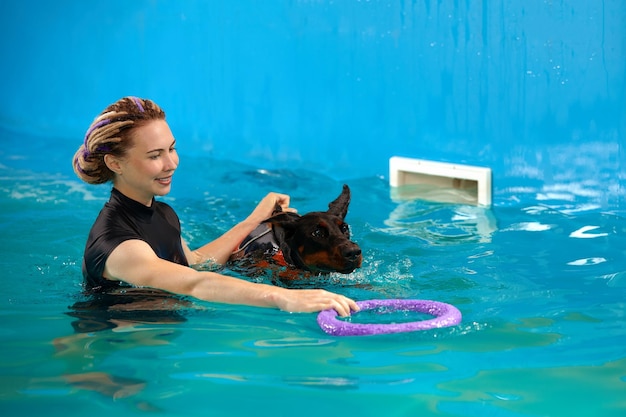 ライフジャケットを着た犬がコーチと一緒にプールで泳ぐ ペットのリハビリ ハイドロセラピーの回復訓練 予防 ペットの健康管理