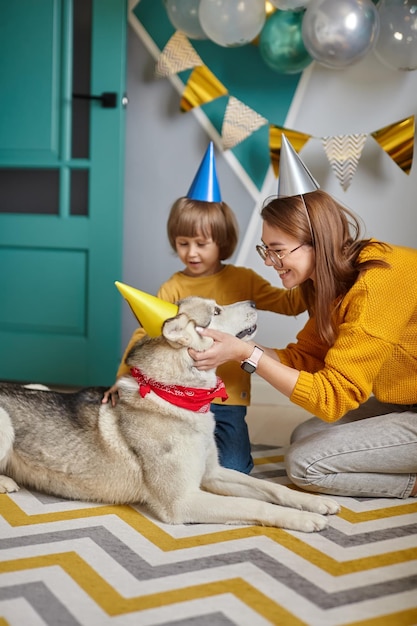 Foto la madre e il bambino della famiglia di compleanno dell'animale domestico del cane mettono un cappello festivo sulla testa del cane congratulandosi per il compleanno